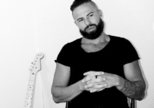 A person with a beard sits casually with hands clasped, next to an electric guitar leaning against the wall in a black and white setting.