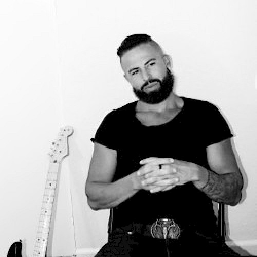 A person with a beard sits casually with hands clasped, next to an electric guitar leaning against the wall in a black and white setting.