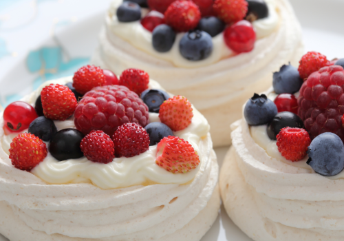 The image shows three meringue nests topped with whipped cream and assorted fresh berries, including raspberries, strawberries, and blueberries.