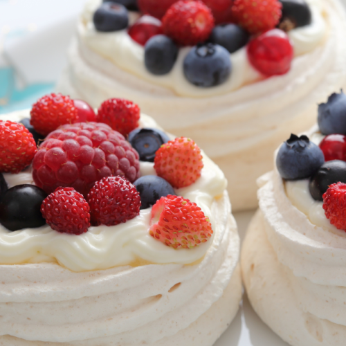 The image shows three meringue nests topped with whipped cream and assorted fresh berries, including raspberries, strawberries, and blueberries.