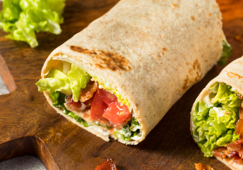 The image shows two wraps filled with lettuce, tomato, and what appears to be bacon, placed on a wooden cutting board with a few pieces on the board.