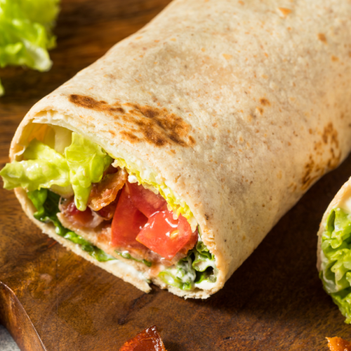 The image shows two wraps filled with lettuce, tomato, and what appears to be bacon, placed on a wooden cutting board with a few pieces on the board.