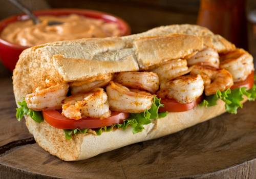 The image showcases a sandwich filled with shrimp, lettuce, and tomato on a wooden board, with a bowl of sauce in the background.