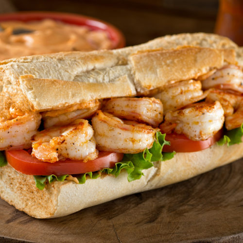 The image showcases a sandwich filled with shrimp, lettuce, and tomato on a wooden board, with a bowl of sauce in the background.