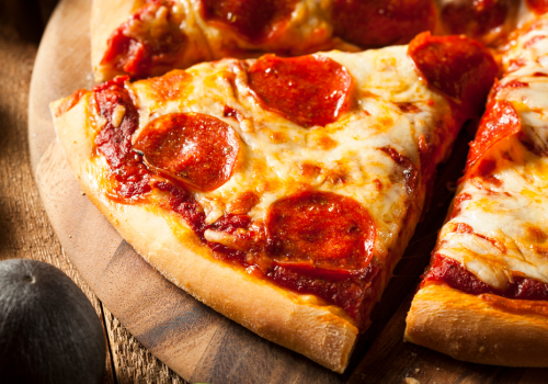 The image shows a close-up of pepperoni pizza slices on a wooden pizza board with some green leaves and a black object on the side.