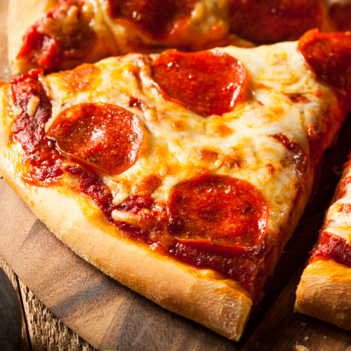 The image shows a close-up of pepperoni pizza slices on a wooden pizza board with some green leaves and a black object on the side.