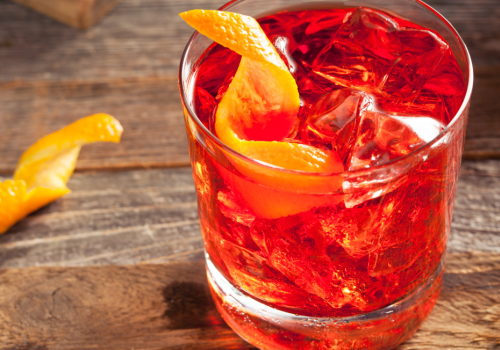 A glass filled with a reddish drink, ice cubes, and an orange peel garnish on a wooden surface. Another orange peel sits next to the glass.