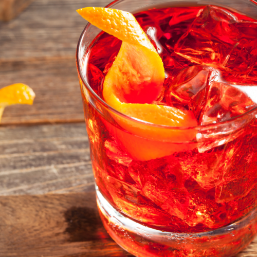 A glass filled with a reddish drink, ice cubes, and an orange peel garnish on a wooden surface. Another orange peel sits next to the glass.