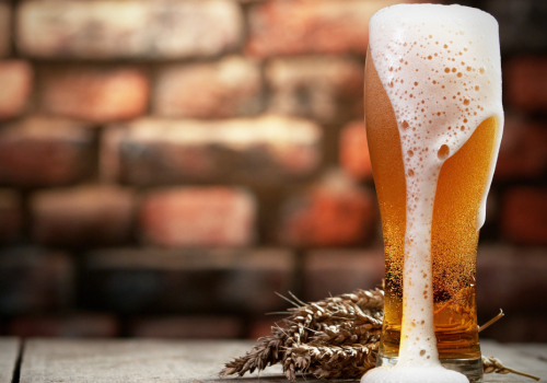 A tall glass of beer with foam overflowing, placed on a wooden surface with wheat sprigs in front of a blurred brick wall background.