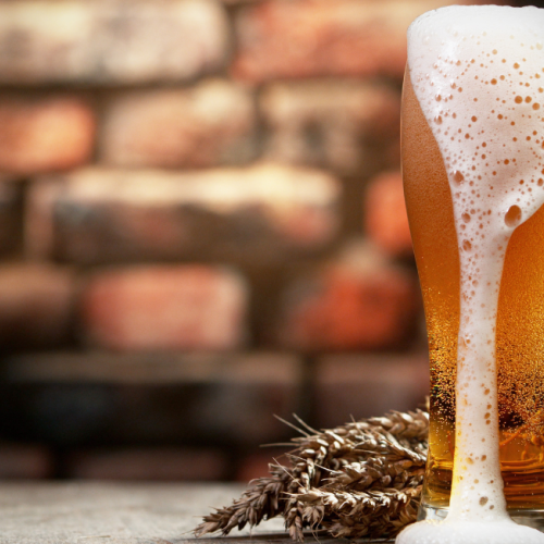 A tall glass of beer with foam overflowing, placed on a wooden surface with wheat sprigs in front of a blurred brick wall background.