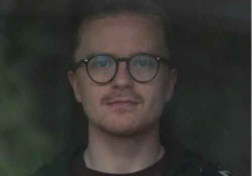 A person with glasses and facial hair, wearing a patterned jacket is looking towards the camera with a neutral expression, taken through a reflective window.