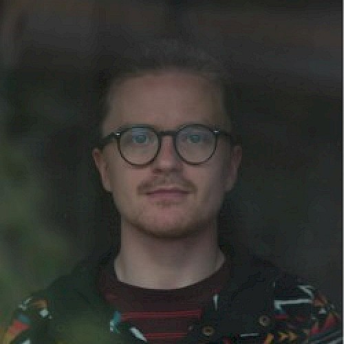 A person with glasses and facial hair, wearing a patterned jacket is looking towards the camera with a neutral expression, taken through a reflective window.