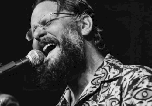 A person with glasses and a beard is passionately singing into a microphone, wearing a patterned shirt. The image is in black and white.