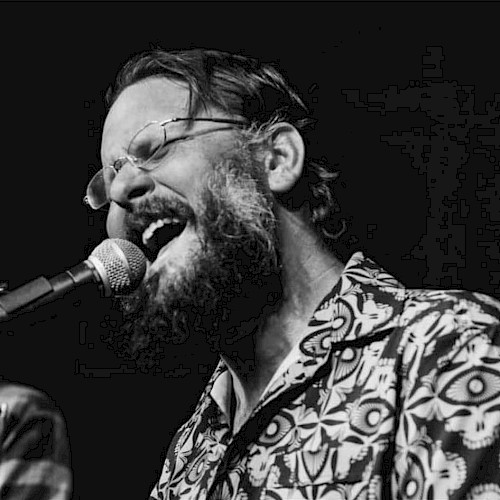 A person with glasses and a beard is passionately singing into a microphone, wearing a patterned shirt. The image is in black and white.