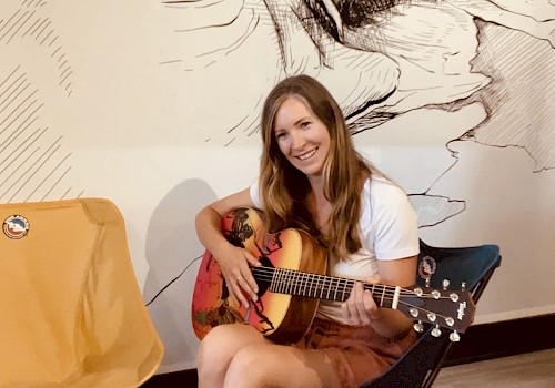 A woman is sitting on a chair, holding a guitar and smiling. There is an empty chair and a woven basket beside her. A mural is in the background.