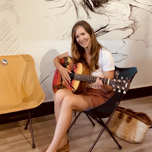 A woman is sitting on a chair, holding a guitar and smiling. There is an empty chair and a woven basket beside her. A mural is in the background.