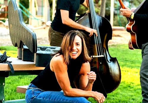 A woman in jeans and a black top sits on a bench outdoors, smiling, with musicians playing string instruments in the background.