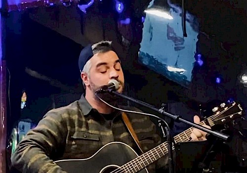 A person wearing a cap plays an acoustic guitar and sings into a microphone on stage, with blue and purple lighting in the background.