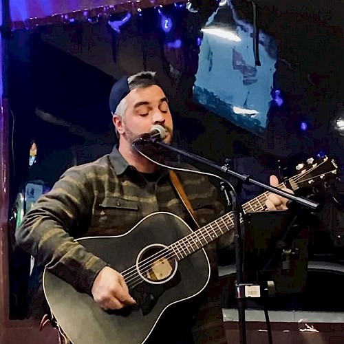 A person wearing a cap plays an acoustic guitar and sings into a microphone on stage, with blue and purple lighting in the background.