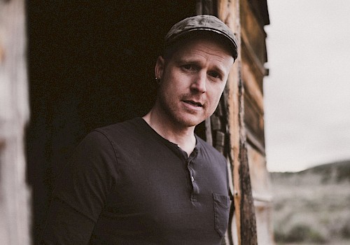 A person in a black shirt and cap stands in a rustic wooden structure, looking outside. The background features an outdoor setting.