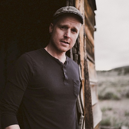 A person in a black shirt and cap stands in a rustic wooden structure, looking outside. The background features an outdoor setting.