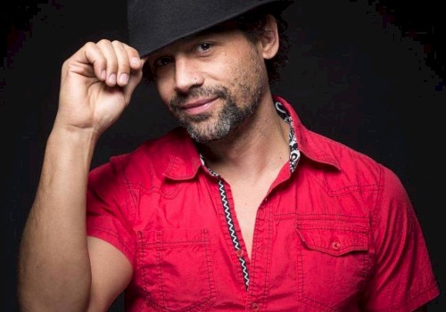 A man in a pink shirt and black hat posing against a dark background while tilting his hat with his right hand.