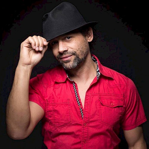 A man in a pink shirt and black hat posing against a dark background while tilting his hat with his right hand.