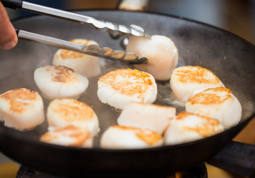 A person is using tongs to cook several seared scallops in a frying pan.