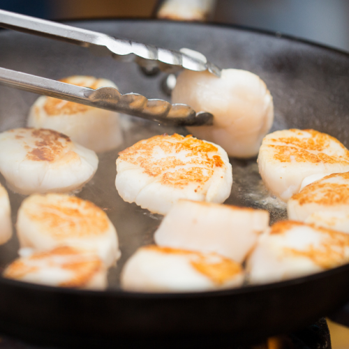 A person is using tongs to cook several seared scallops in a frying pan.