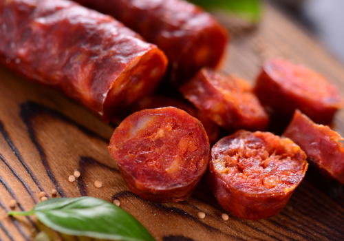 The image shows sliced pieces of cured sausage on a wooden surface, alongside some green leaves, possibly herbs.