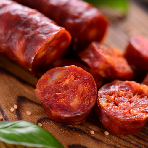 The image shows sliced pieces of cured sausage on a wooden surface, alongside some green leaves, possibly herbs.