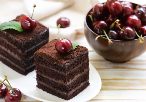 The image shows two pieces of layered chocolate cake topped with cherries on a white plate and a wooden bowl filled with fresh cherries in the background.