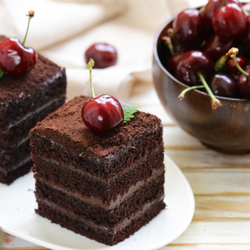 The image shows two pieces of layered chocolate cake topped with cherries on a white plate and a wooden bowl filled with fresh cherries in the background.