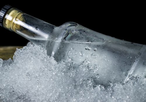 The image shows a clear glass bottle with a black and gold cap, partially submerged in crushed ice, with a dark background.