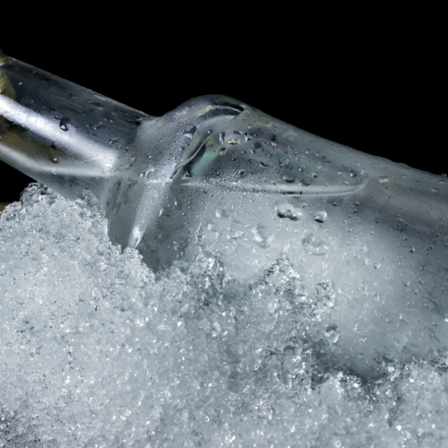 The image shows a clear glass bottle with a black and gold cap, partially submerged in crushed ice, with a dark background.