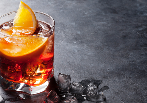 A glass filled with a dark-colored beverage, ice cubes, and garnished with an orange slice sits on a dark, textured surface with scattered ice cubes.