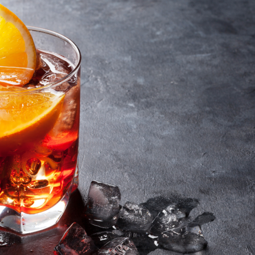 A glass filled with a dark-colored beverage, ice cubes, and garnished with an orange slice sits on a dark, textured surface with scattered ice cubes.