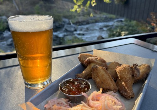 A glass of beer sits beside a tray of fried snacks, sauce, and pickled vegetables on an outdoor table with a scenic view of nature.