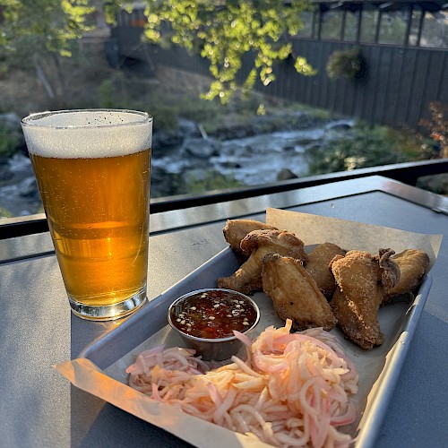 A glass of beer sits beside a tray of fried snacks, sauce, and pickled vegetables on an outdoor table with a scenic view of nature.