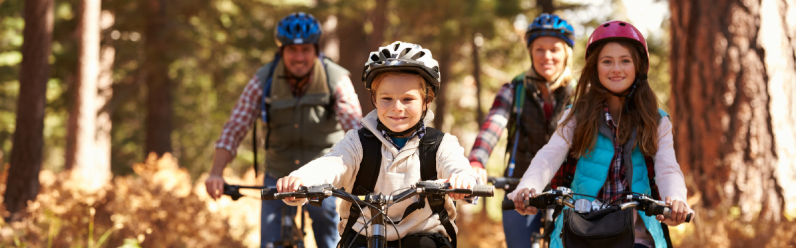 A group of four people, two adults and two children, riding bicycles on a forest trail, all wearing helmets and smiling.