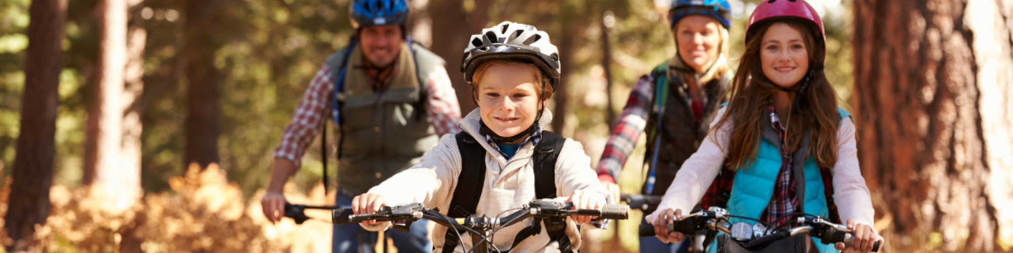 A group of four people, two adults and two children, riding bicycles on a forest trail, all wearing helmets and smiling.