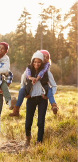 A family of four enjoys a piggyback ride together in a serene outdoor setting, with trees and sunlight in the background.