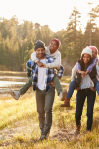 A family of four enjoys a piggyback ride together in a serene outdoor setting, with trees and sunlight in the background.