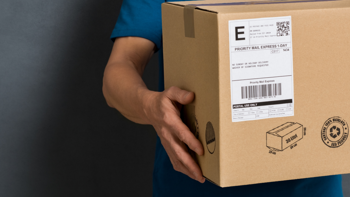 A person is holding a cardboard box with a shipping label and recycling symbol, suggesting it’s ready for delivery.