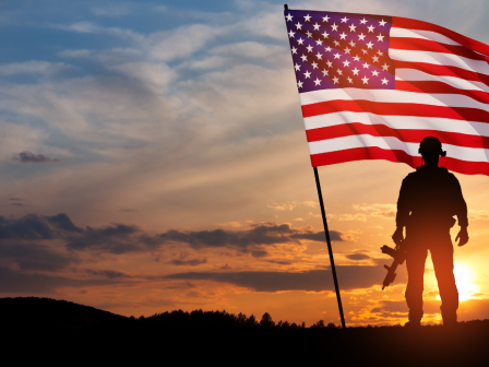 A silhouetted soldier stands with a rifle and a large American flag against a dramatic sunset sky.