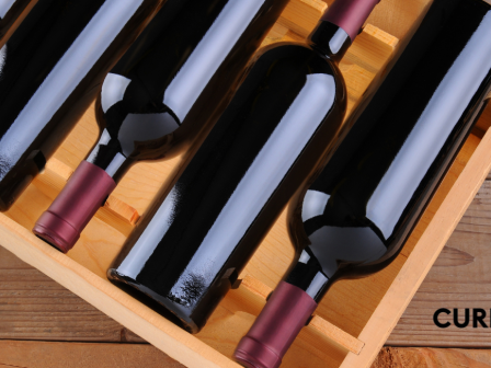 The image shows five wine bottles with red foil caps in a wooden box, placed on a wooden surface. The word 