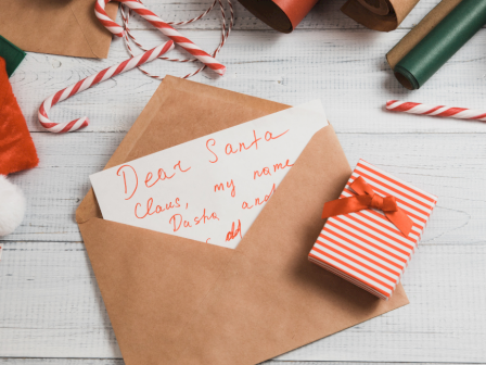 A letter to Santa, wrapped present, candy canes, and holiday decorations are arranged on a white wooden surface.