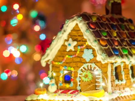 A decorated gingerbread house with icing and colorful candies, set against a background of festive, blurred lights.