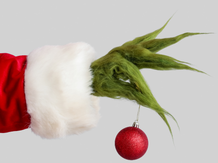 A green furry hand in a red Santa sleeve holds a red Christmas ornament against a plain background.