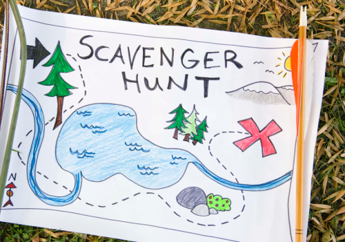 A hand-drawn scavenger hunt map on grass features a lake, trees, a mountain, and a red X, suggesting a treasure location.